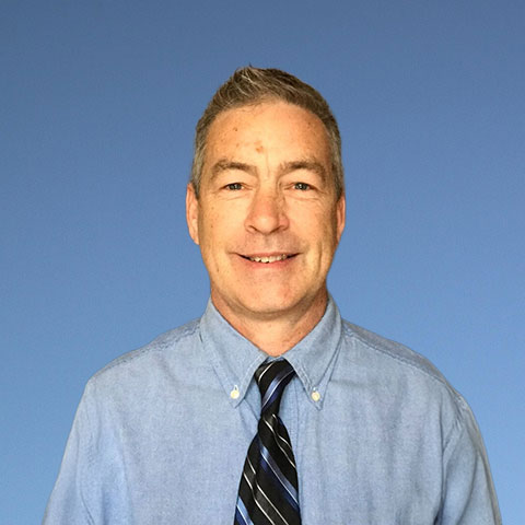A headshot of Jeffrey Buck with short, styled hair. He is wearing a light blue button-down shirt with a dark blue, light blue, and black striped tie. He stands in front of a rustic wooden plank wall, facing the viewer directly.